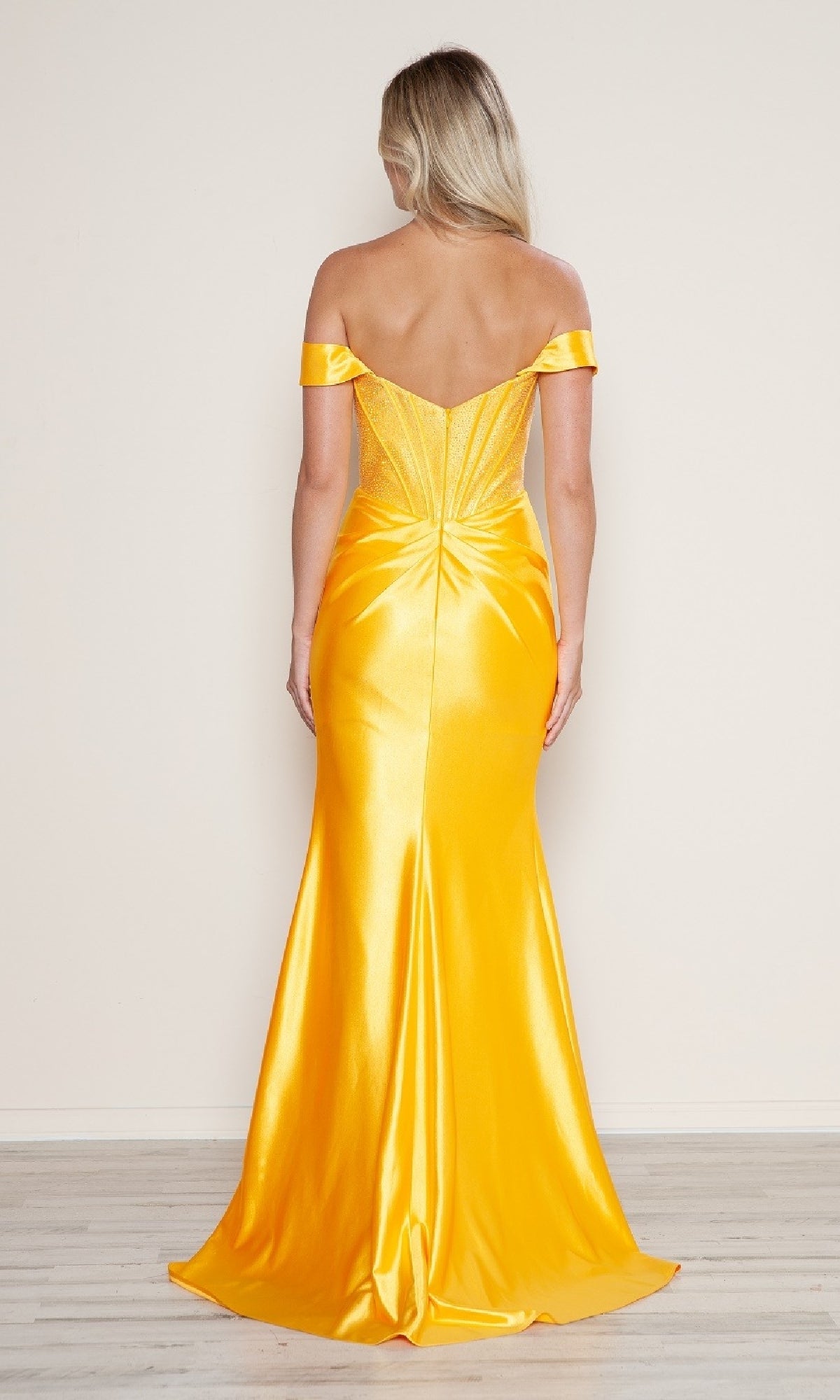 A woman with long blonde hair is seen from the back wearing the Off-Shoulder Long Corset Prom Dress: Poly USA 9656, a fitted yellow satin gown with a flared skirt and beaded corset details, set against a plain light background.